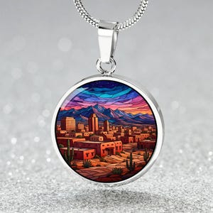 May include: A silver pendant necklace featuring a round charm with a colorful desert landscape design. The charm depicts a city with mountains, buildings, and cacti under a vibrant sunset sky. The necklace has a silver chain.