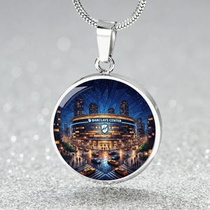 May include: A silver-toned pendant necklace featuring a round charm with a colorful illustration of the Barclays Center at night. The image includes the text "BARCLAYS CENTER" and a logo. The necklace has a silver chain.