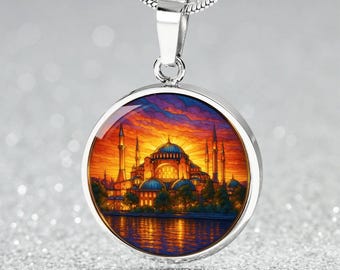 Puede incluir: Un collar con colgante plateado con un dije redondo que representa una colorida ilustración de un edificio con múltiples minaretes y cúpulas, sobre un cielo al atardecer. El colgante cuelga de una cadena plateada.