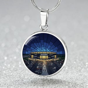 May include: A silver-toned pendant necklace featuring a round charm with a detailed illustration of UBS Arena under a night sky. The arena is lit up, with a parking lot and people visible. The necklace hangs on a silver chain.