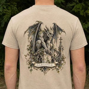 May include: A sand-coloured t-shirt featuring a detailed graphic on the back. The print depicts a winged gargoyle perched atop a stone structure with ivy accents. Below the gargoyle, a banner displays the text GARGOYLE in capital letters.