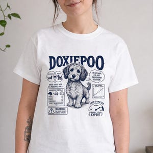 May include: A white t-shirt featuring a dark blue graphic of a Doxiepoo dog. The design includes text reading DOXIEPOO, a Venn diagram, a formula, a warning sign, a bark volume graph, and a sass-o-meter gauge with the text CURRENT LEVEL: EXPERT.