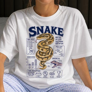 Könnte beinhalten: Ein weißes T-Shirt mit einer zentralen Grafik einer geringelten braunen und beigen Schlange. Das Design enthält den Text "SNAKE", "MOTTO: DO LESS. HISS MORE. NAP OFTEN.", "SNEK FACTS", "VENOM OUTPUT", "SNAKE SHAPE EXPLAINED", "ANATOMY 101" und "CARE INSTRUCTIONS".