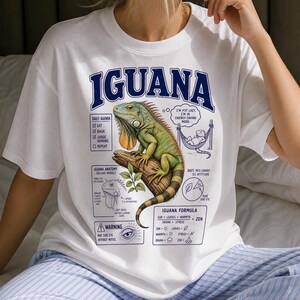 Op de afbeelding: Een wit t-shirt met een grote afbeelding van een groene leguaan op een tak. Het ontwerp bevat teksten zoals IGUANA, DAILY AGENDA, IGUANA ANATOMY en IGUANA FORMULA, samen met een kleine illustratie van een leguaan in een hangmat en een cirkeldiagram met het label DIET.