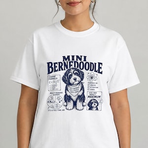 May include: A white t-shirt featuring a dark blue graphic of a Mini Bernedoodle dog wearing a bandana. The design includes text such as "MINI BERNEDOODLE," a "CANINE FORMULA" chart, a "MULTITASKING EXPERT" pie chart, and "POWERED BY" checkboxes.