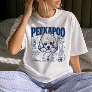 May include: A white t-shirt featuring a blue graphic of a fluffy dog with the text PEEKAPOO. The design includes a stealth equation, a gauge labeled ninja level, and text bubbles reading I SEE YOU. YOU JUST DON'T SEE ME and THEY THINK I'M CUTE.