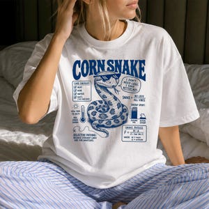 Op de afbeelding: Een wit t-shirt met een blauwe afbeelding van een korenslang met een gepixelde zonnebril. Het ontwerp bevat tekst zoals CORN SNAKE, een verzorgingschecklist, een thermometer-icoon en een natuurkundige vergelijking in donkerblauwe inkt.