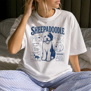 Op de afbeelding: Een wit t-shirt met een blauwe afbeelding van een Sheepadoodle-hond. Het ontwerp bevat de tekst SHEEPADOODLE, een wiskundige vergelijking, een vaardighedengrafiek en een waarschuwingsvak. De illustratie toont een pluizige hond met donkere oren.