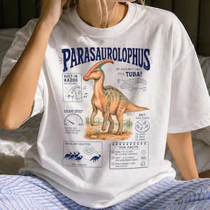 May include: A white t-shirt featuring a graphic of a Parasaurolophus dinosaur with text labels including "Built-in Kazoo," "Not just a pretty crest, it's a tuba!," "Escape speed," "Diet: 100% plants," "Social but selective," and a "Fun facts" list.