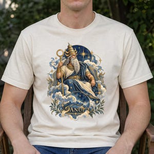 May include: A cream-colored t-shirt featuring a graphic illustration of a bearded deity wearing a horned crown and robes, holding a staff. The figure is surrounded by clouds, stars, and bull motifs, with the text ANU centered at the bottom.
