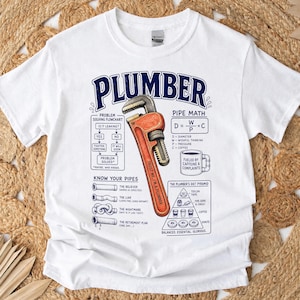 May include: A white short-sleeve t-shirt featuring a graphic print with the word PLUMBER in blue. The design includes a red pipe wrench, a problem-solving flowchart, a pipe math equation, a coffee mug, pipe types, and a plumber's diet pyramid.