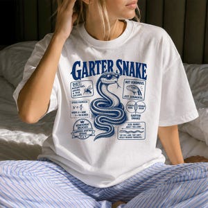 Puede incluir: Una camiseta blanca de manga corta con un gráfico azul de una culebra de liga con gafas de sol. El diseño incluye textos como Garter Snake, Diet: Mosquitoes, Worms, & Your Excuses, Speed Formula, Not Venomous, Just Judgmental y Size Range.