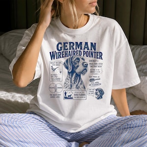 Peut inclure: Un t-shirt blanc à manches courtes avec un motif bleu représentant un braque allemand à poil dur. Le design inclut des textes comme "Built-in GPS", "Energy = Walk + Run + Fetch", "Coat made of: Steel Wool" et "Official Job Title: Bird Flusher."