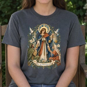 Op de afbeelding: Een antracietgrijs t-shirt met een grafische illustratie van de tarotkaart Zes van Pentakels. Het ontwerp toont een centrale figuur met een weegschaal, geflankeerd door twee knielende personen, omringd door witte lelies, duiven en een banner met de tekst "SIX OF PENTACLES" onderaan.