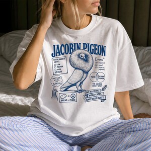May include: A white t-shirt featuring a blue graphic of a Jacobin pigeon with a large ruff. The design includes text such as "Jacobin Pigeon," "Social Anxiety Level: Ruff," "E=mc2," "Bread = Peace," and "Not all heroes wear capes. Some wear ruffs."