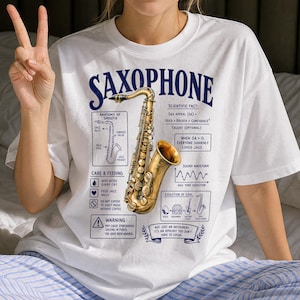 Op de afbeelding: Een wit T-shirt met korte mouwen en een grote afbeelding van een gouden saxofoon. Het ontwerp bevat teksten zoals SAXOPHONE, ANATOMY OF SMOOTH, SCIENTIFIC FACT en diverse diagrammen, grafieken en humoristische waarschuwingen in donkerblauw.