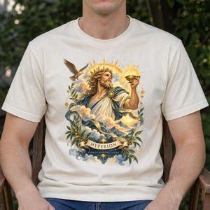 May include: A cream-coloured t-shirt featuring a graphic illustration of a bearded figure with a crown, holding a glowing orb. The design includes a flying eagle, clouds, mountains, and a banner with the text HYPERION TITAN OF HEAVENLY LIGHT.