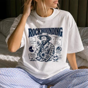 以下が含まれることがあります： ROCKHOUNDINGという文字がプリントされたダークブルーのグラフィックが特徴の白いTシャツ。帽子をかぶった髭の人物が石を持っている様子、チェックリスト、円グラフ、数式、CAUTIONと書かれた看板、サングラスをかけたキャラクター化された石が描かれています。