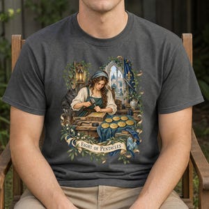 Op de afbeelding: Een antracietgrijs t-shirt met een gedetailleerde afbeelding van een persoon die aan een houten werkbank werkt en gouden munten bewerkt. De scène bevat een zwarte kat, een lantaarn en een banner met de tekst EIGHT OF PENTACLES in zwart.
