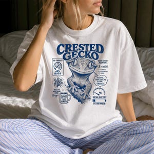 Può includere: Una t-shirt bianca con una grafica blu di un geco crestato. Il design include il testo "Crested Gecko", "No thoughts only bugs", "Built-in wall climb mode", "Humans are just bug servers" e "Feed me, then leave me alone, forever."