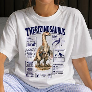 May include: A white t-shirt featuring a graphic of a Therizinosaurus dinosaur with text detailing its diet, claw size of 1.0m, and a damage chart. The design includes humorous labels like "I'm not aggressive, I'm just passionate about pruning" in blue ink.
