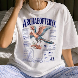 Può includere: Una t-shirt bianca con la grafica di un dinosauro Archaeopteryx con piume blu e arancioni. Il design include testo come ARCHAEOPTERYX, un piano di volo, un diagramma multi-strumento e una lista di controllo dell'identità con caselle.