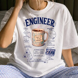 Puede incluir: Una camiseta blanca de manga corta con un estampado gráfico titulado ENGINEER. El diseño incluye una taza de café con una nota adhesiva que dice TRUST ME, I'M AN ENGINEER, un diagrama de flujo, un esquema cerebral y textos técnicos.