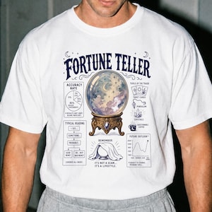 May include: A white short-sleeve t-shirt featuring a graphic print titled FORTUNE TELLER. The design includes a crystal ball on a stand, a pie chart labeled ACCURACY RATE, tarot cards, a pendulum, crystals, a coffee mug, and a flow chart.
