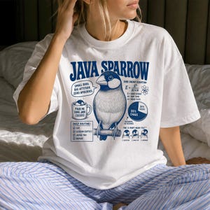 Può includere: Una t-shirt bianca a maniche corte con una grafica blu di un passero di Giava. Il design include il testo Java Sparrow, una lista di controllo della routine quotidiana, un grafico a torta con 80% chaos e un'equazione energetica aviaria.