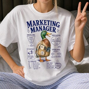 Könnte beinhalten: Ein weißes T-Shirt mit einer Grafik einer Stockente, die eine Umhängetasche trägt. Der Text lautet Marketing Manager, Marketing Funnel, Budget Allocation, My Superpower, Campaign Process, KPI Translator und Daily Goals mit verschiedenen Symbolen.