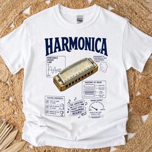 Puede incluir: Una camiseta blanca con un gráfico de una armónica y el texto HARMONICA, PORTABLE MOOD LAB, ANATOMY OF NOISE, PLAYING HARMONICA, THE ORIGINAL POCKET SIZED REGRET y SOUND OUTPUT con varios diagramas y notas musicales impresos en azul oscuro.