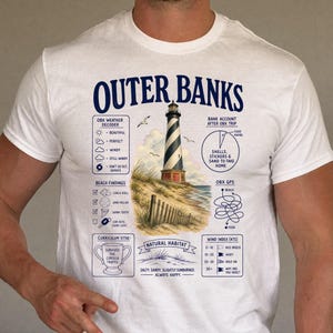 Pode incluir: Uma t-shirt branca de manga curta com uma impressão gráfica intitulada Outer Banks. O design inclui uma ilustração de um farol, um descodificador meteorológico, uma lista de achados de praia, um gráfico circular, um labirinto e texto.