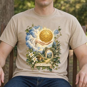 Può includere: Una t-shirt beige con una stampa grafica di una mano che emerge dalle nuvole tenendo una moneta pentacolo dorata. Sotto, un sentiero da giardino conduce a un arco circondato da gigli bianchi e verde. Il testo ACE OF PENTACLES è stampato in basso.