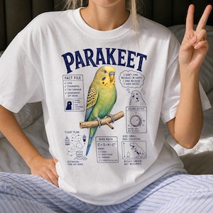Könnte beinhalten: Ein weißes T-Shirt mit einer zentralen Abbildung eines grün-gelben Wellensittichs auf einem Ast. Die Grafik enthält Texte wie PARAKEET, FACT FILE, BIRB MATH sowie verschiedene Diagramme, die das Verhalten des Vogels beschreiben.