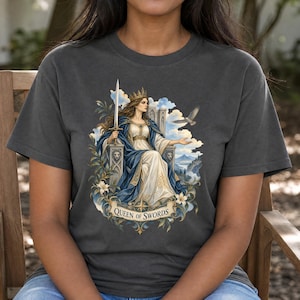 May include: A charcoal grey t-shirt featuring a graphic illustration of a queen sitting on a throne, holding a sword, and wearing a crown. The design includes a bird, clouds, and foliage, with a banner at the bottom reading Queen of Swords.