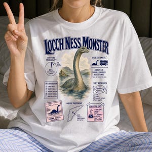 May include: A white t-shirt featuring a graphic of the Loch Ness Monster with text that reads Loch Ness Monster, Spotting Probability, Size Estimate, Field Identification, Diet Research, Sonar Photo, Habitat Preference, and Scientific Fact.