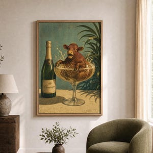 May include: A framed wall art print featuring a brown cow sitting inside a large glass of champagne, with a green champagne bottle standing next to it. The background shows dark green palm leaves against a muted teal wall, with a light tan surface.