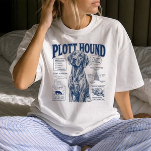 Puede incluir: Una camiseta blanca con un estampado gráfico azul de un perro Plott Hound. El diseño incluye el texto "PLOTT HOUND", una lista de verificación "Bred For", una fórmula matemática "Scent Algorithm", un gráfico de radar de habilidades y un gráfico "Trash Panda".