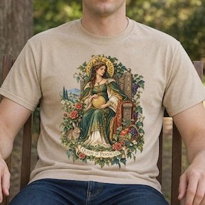 May include: A beige t-shirt featuring a graphic illustration of a woman in a green gown sitting on a throne, holding a gold pentacle. The design includes a rabbit, red roses, grapes, and a banner with the text Queen of Pentacles.