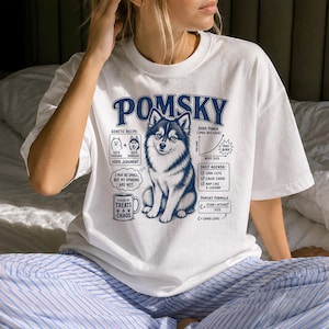 Pode incluir: Uma t-shirt branca com um gráfico azul escuro de um cão Pomsky. O design inclui o texto POMSKY, um diagrama de receita genética, um gráfico de potência de latido, uma agenda diária, uma caneca com a etiqueta FUELED BY TREATS & CHAOS e uma fórmula.