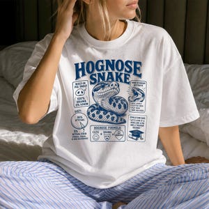 May include: A white t-shirt featuring a blue graphic of a hognose snake with text including HOGNOSE SNAKE, BUILT-IN PIG SNOUT, 100% DRAMA 0% VENOM, DIET: 95% ATTITUDE, HOGNOSE FORMULA, WHEN IN DANGER: ACTIVATE, and EVOLUTION SAID: LET'S GIVE IT A NOSE.