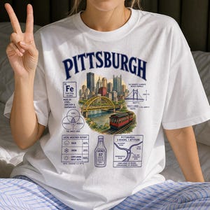 May include: A white t-shirt featuring a graphic print of the Pittsburgh skyline, a yellow bridge, and a red trolley. The design includes text elements like 'PITTSBURGH', a periodic table style 'Fe' box, a weather report, a Heinz 57 bottle, and a river map.