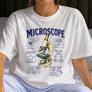 May include: A white t-shirt featuring a central graphic of a microscope surrounded by scientific diagrams and text including "MICROSCOPE," "CELLFIE KING," "MAGNIFICATION BOOST MODE," "SAMPLE TODAY, BRAG FOREVER," "WHAT I SEE," and "RESOLUTION > OPINIONS."