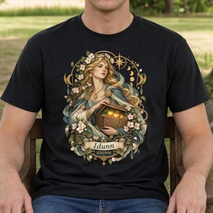 May include: A black t-shirt featuring a graphic illustration of a woman with long blonde hair adorned with flowers, holding a wooden chest filled with golden apples. The design includes celestial moon phases, a star, and a banner with the text Idunn.