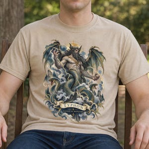 May include: A beige short-sleeve t-shirt featuring a detailed graphic print of the mythological figure Typhon. The design depicts a winged, bearded humanoid with a serpentine lower body, surrounded by lightning, clouds, and snakes. Text reads TYPHON FATHER OF MONSTERS.