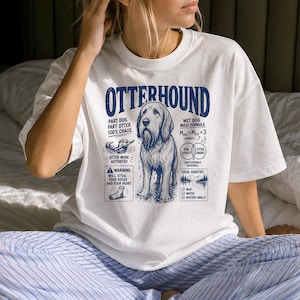 Może przedstawiać: Biały t-shirt z krótkim rękawem z niebieską grafiką psa rasy Otterhound. Tekst zawiera napisy OTTERHOUND, PART DOG. PART OTTER. 100% CHAOS., WET DOG MASS FORMULA, WARNING: WILL STEAL YOUR SOCKS AND YOUR HEART, oraz IDEAL HABITAT: MUD, WATER, MYSTERY SMELLS.