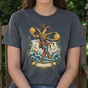 Pode incluir: Uma t-shirt cinza-carvão com uma estampa gráfica de um bobo da corte em roupas azuis e vermelhas equilibrando dois pentáculos dourados conectados por uma fita. A cena inclui ondas do mar, dois veleiros e uma faixa com o texto Two of Pentacles.