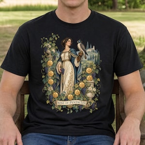 May include: A black t-shirt featuring a graphic illustration of a person in a white dress and blue cloak holding a bunch of grapes, with a falcon perched on their arm. The design includes nine gold pentacles, grapevines, and the text Nine of Pentacles.
