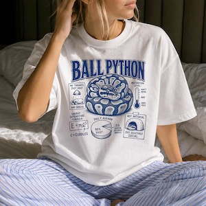 Könnte beinhalten: Ein weißes Kurzarm-T-Shirt mit einer blauen Grafik eines zusammengerollten Königspythons. Das Design enthält die Textbeschriftungen: Ball Python, No Thoughts Just Heat And Snacks, Motivation Level, Energy Expenditure Formula E=mc2, Daily Agenda und Not Antisocial Just Selectively Social.