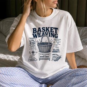 May include: A white t-shirt featuring a dark blue graphic print with the text BASKET WEAVING. The design includes a central woven basket, a coffee mug, a circular weave cycle diagram, and text detailing a woven happiness formula.